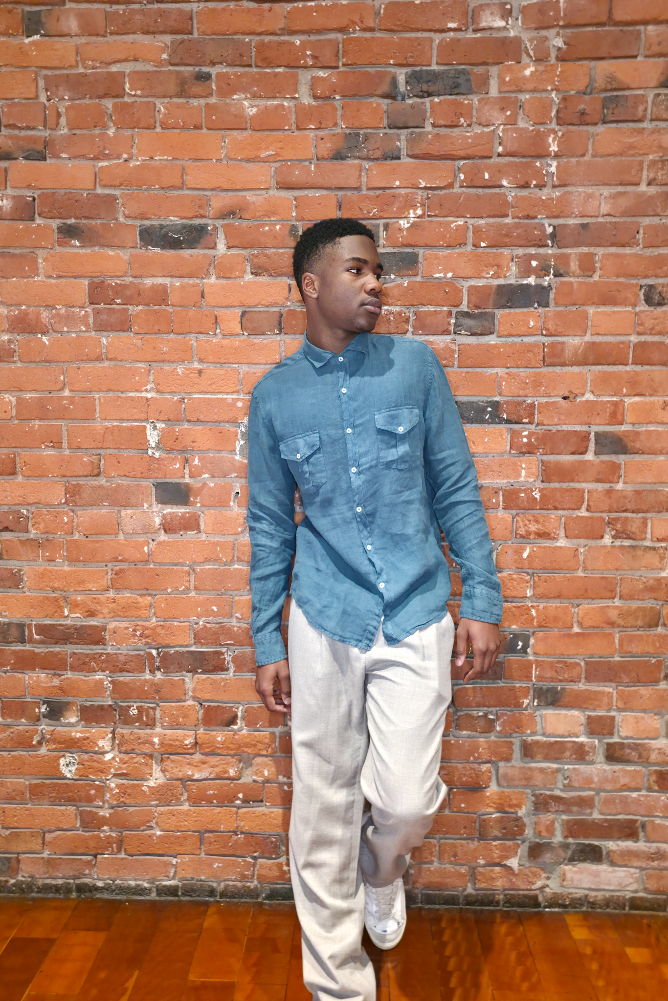 Man wearing a blue shirt and beige pants standing against a brick wall.