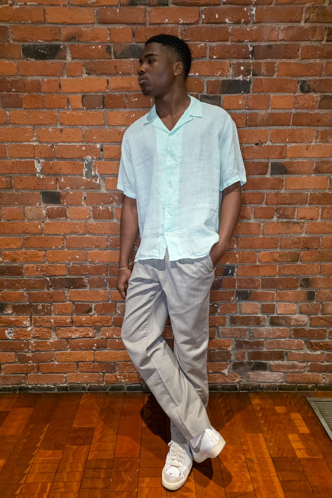 Man standing against a brick wall wearing a light blue shirt and beige pants.