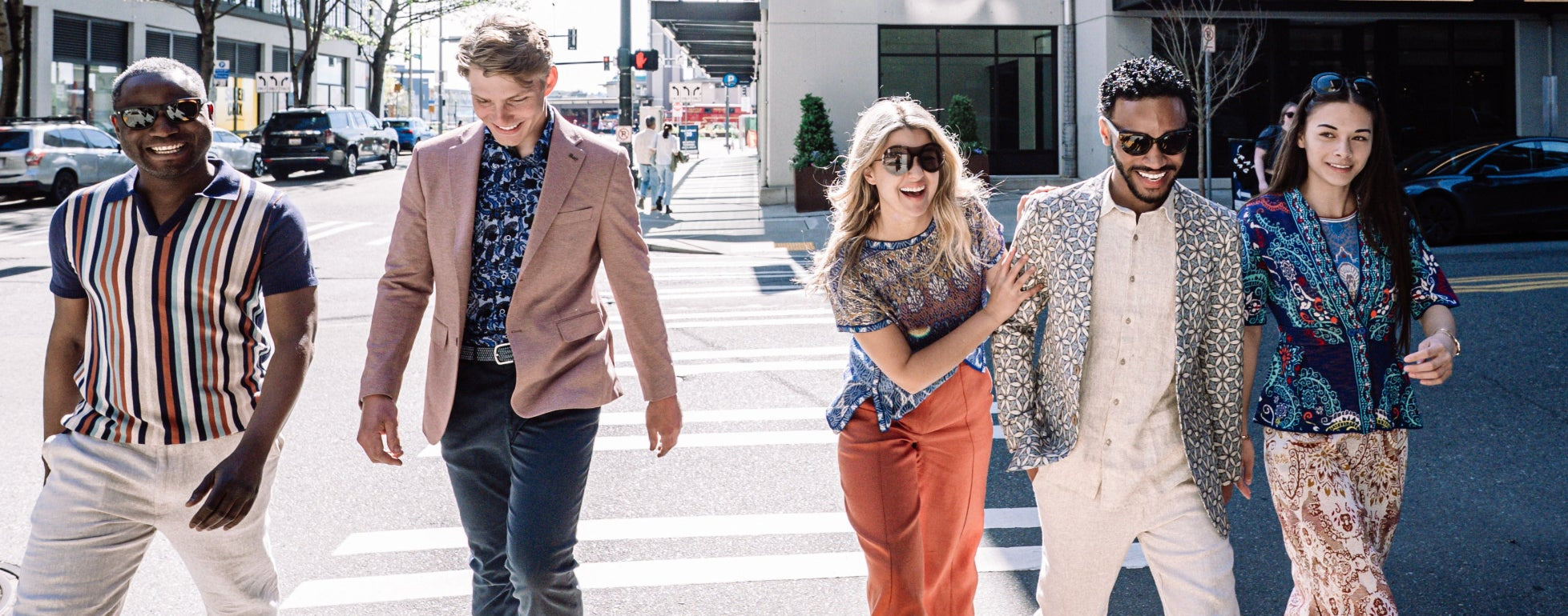 Five stylish people walking across a city crosswalk in bright daylight, each wearing unique fashion looks including patterned shirts, blazers, and wide-leg pants.