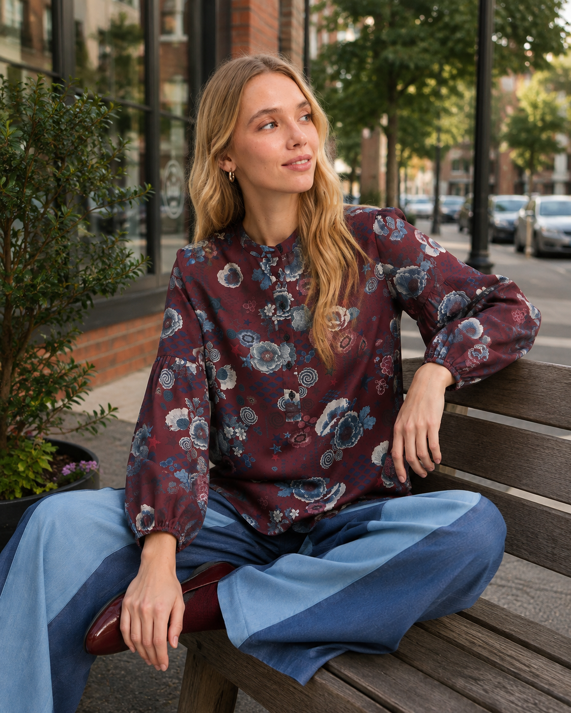 A woman sits on a wooden park bench wearing a long-sleeve high-neck blouse with a burgundy, blue, and gray floral pattern, paired with blue patchwork wide-leg trousers.
