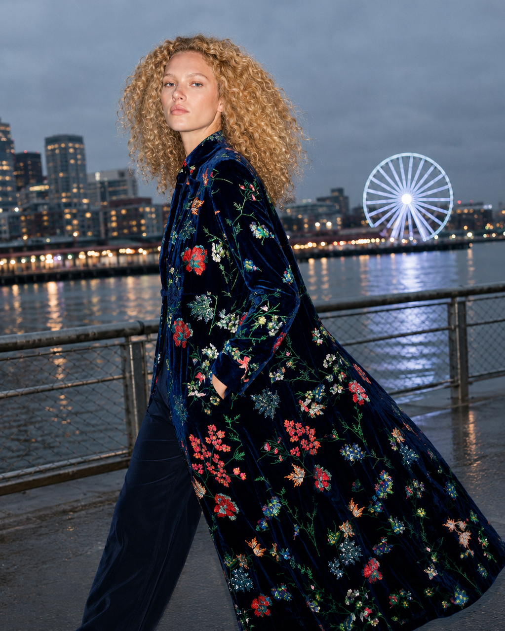 A model with curly blonde hair walks along a waterfront at dusk wearing a long navy velvet coat with a colorful floral print and matching navy trousers. The background features a city skyline and a lit-up Ferris wheel.