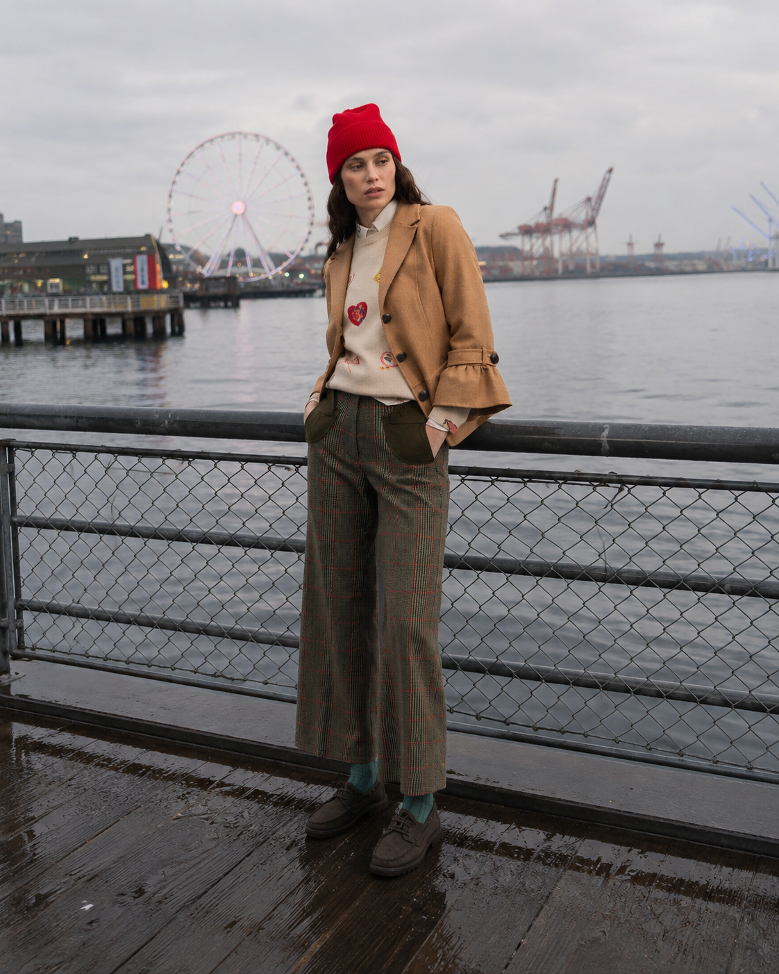 A woman stands on a wet wooden boardwalk by the water, wearing a tan blazer, beige sweater with red heart details, brown plaid wide-leg trousers, a red knit beanie, and brown loafers. A chain-link fence, water, and a Ferris wheel are visible in the background.