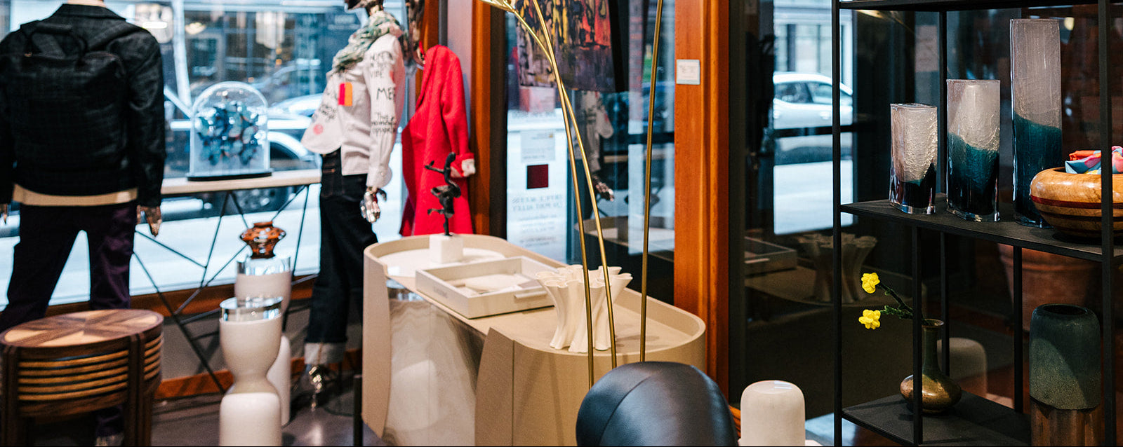 A styled interior boutique scene with mannequins wearing outerwear, a cream console table, tall glass globes hanging from the ceiling, a floor lamp, and shelving displaying vases and decor. No person or animal is present, but this is not a clean isolated product shot; it is a real retail environment, so subject_type is Lifestyle.