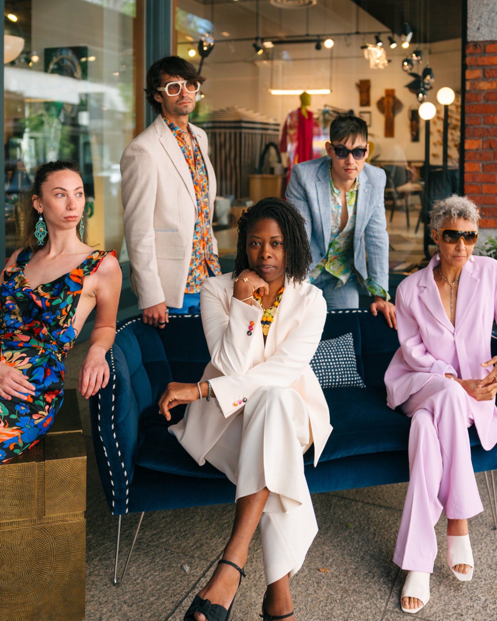 Five models pose together in a stylish indoor boutique setting. A woman in a white blazer sits on a blue velvet sofa, while a man in a cream blazer and a woman in a lavender blazer stand beside her.
