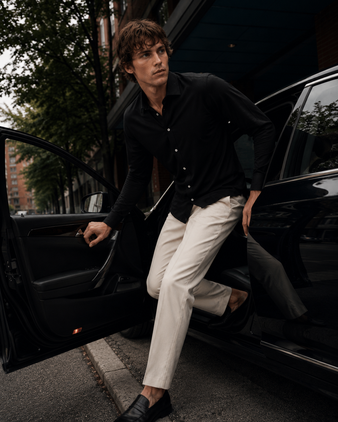 A man in a black button-down shirt and off-white trousers steps out of a black car door on a city street.