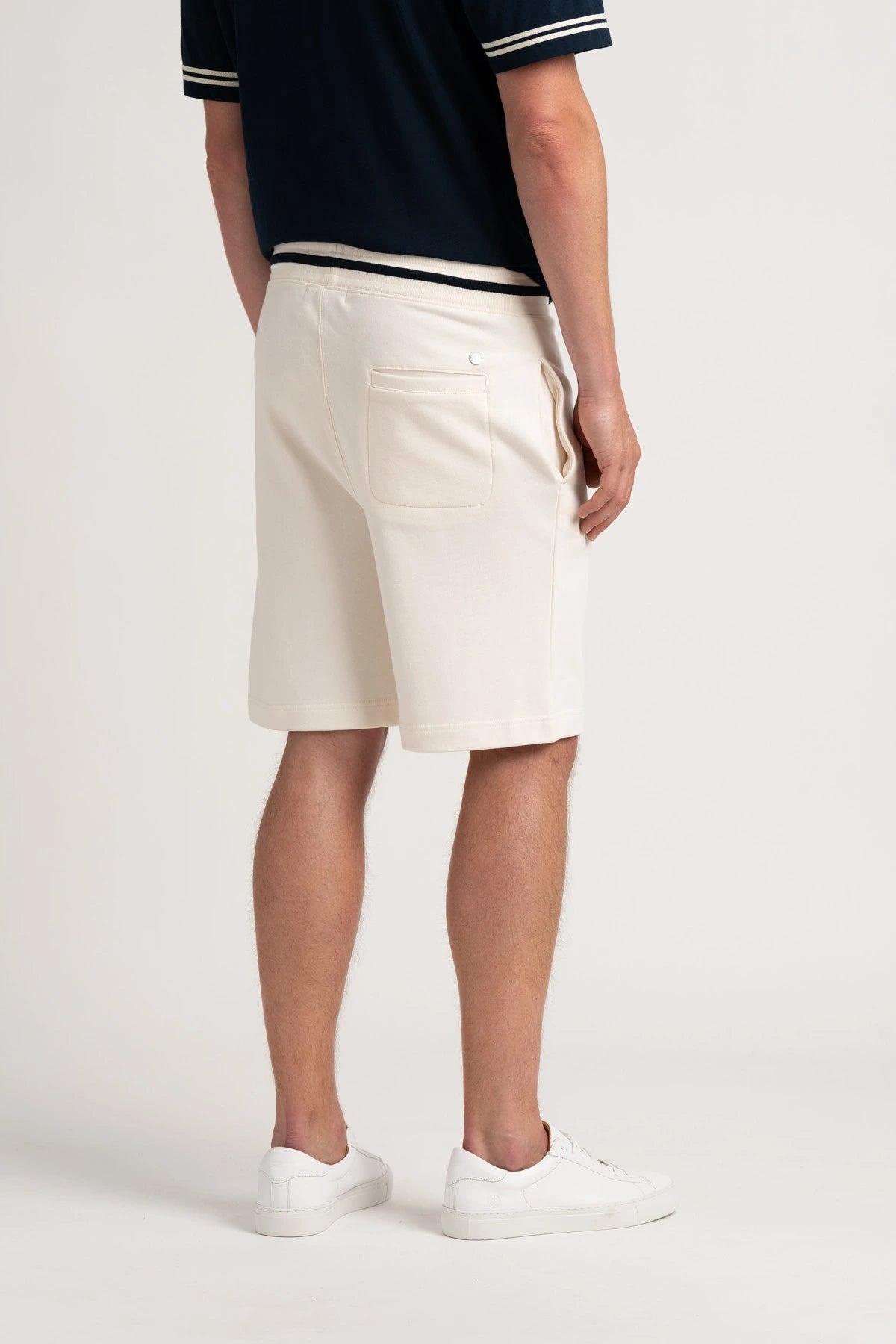 A man shown from the back wearing off-white chino shorts, a navy blue polo shirt with white stripes, and white sneakers, set against a plain light gray studio background.