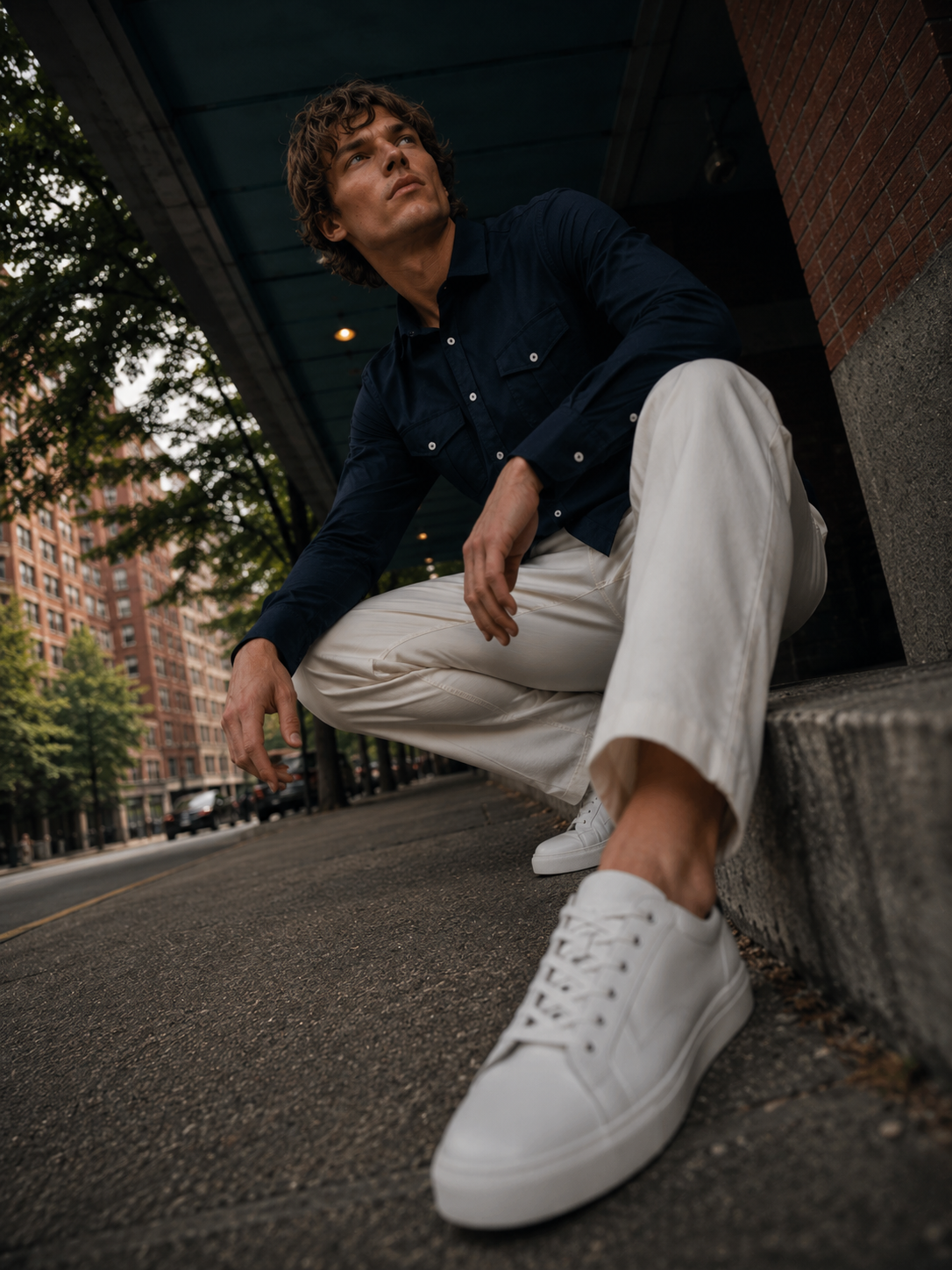 A man crouching on a city sidewalk under a covered walkway, wearing a dark navy long-sleeve button-up shirt, off-white trousers, and white low-top sneakers.