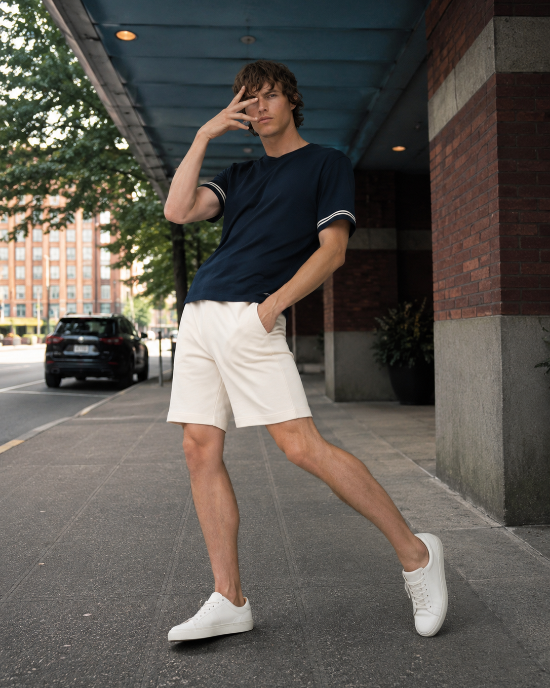 A man posing on an urban sidewalk under a covered walkway, wearing a navy blue t-shirt with white sleeve trim, off-white shorts, and white sneakers.
