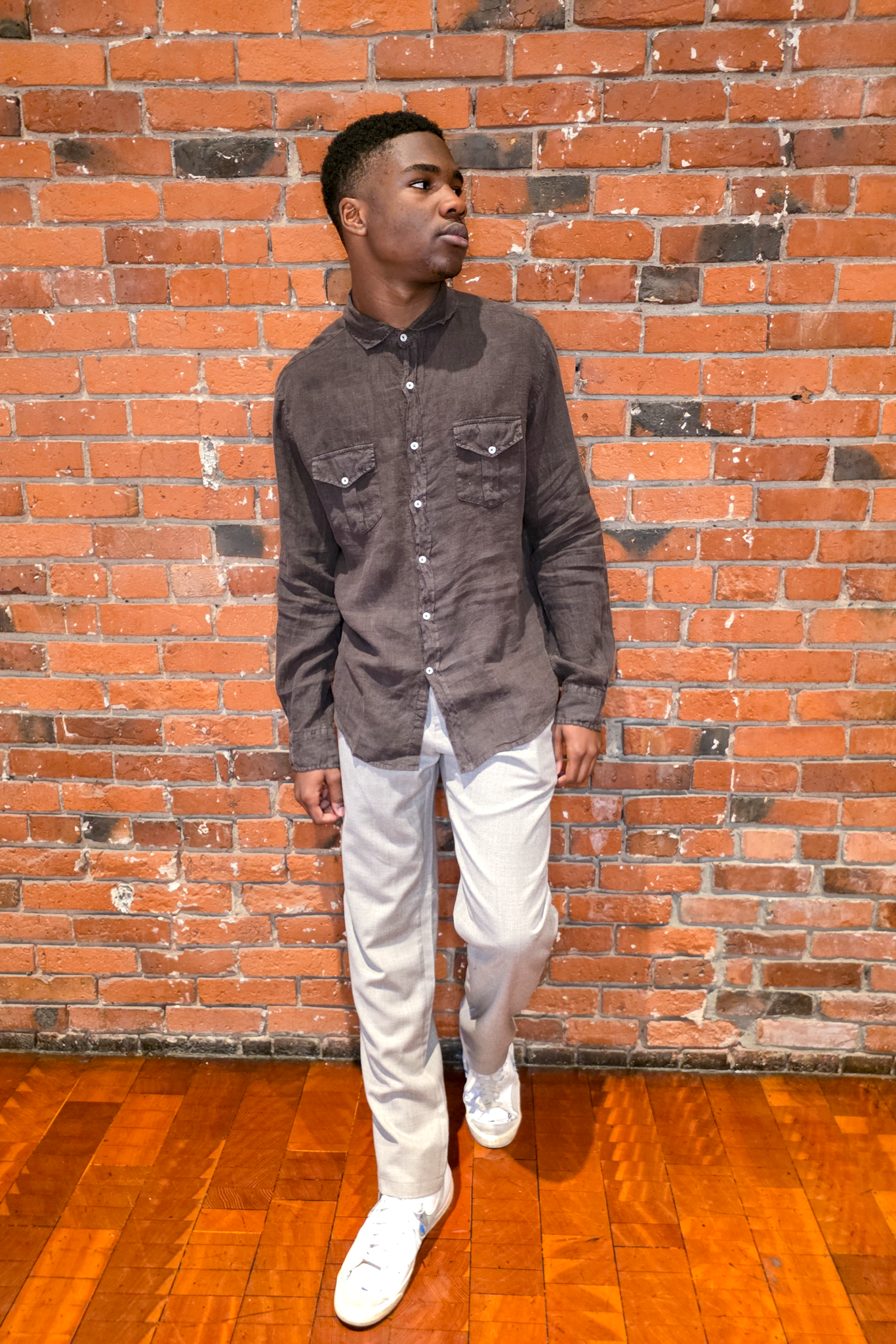 Man wearing a dark gray button-up shirt and white pants standing against a brick wall.