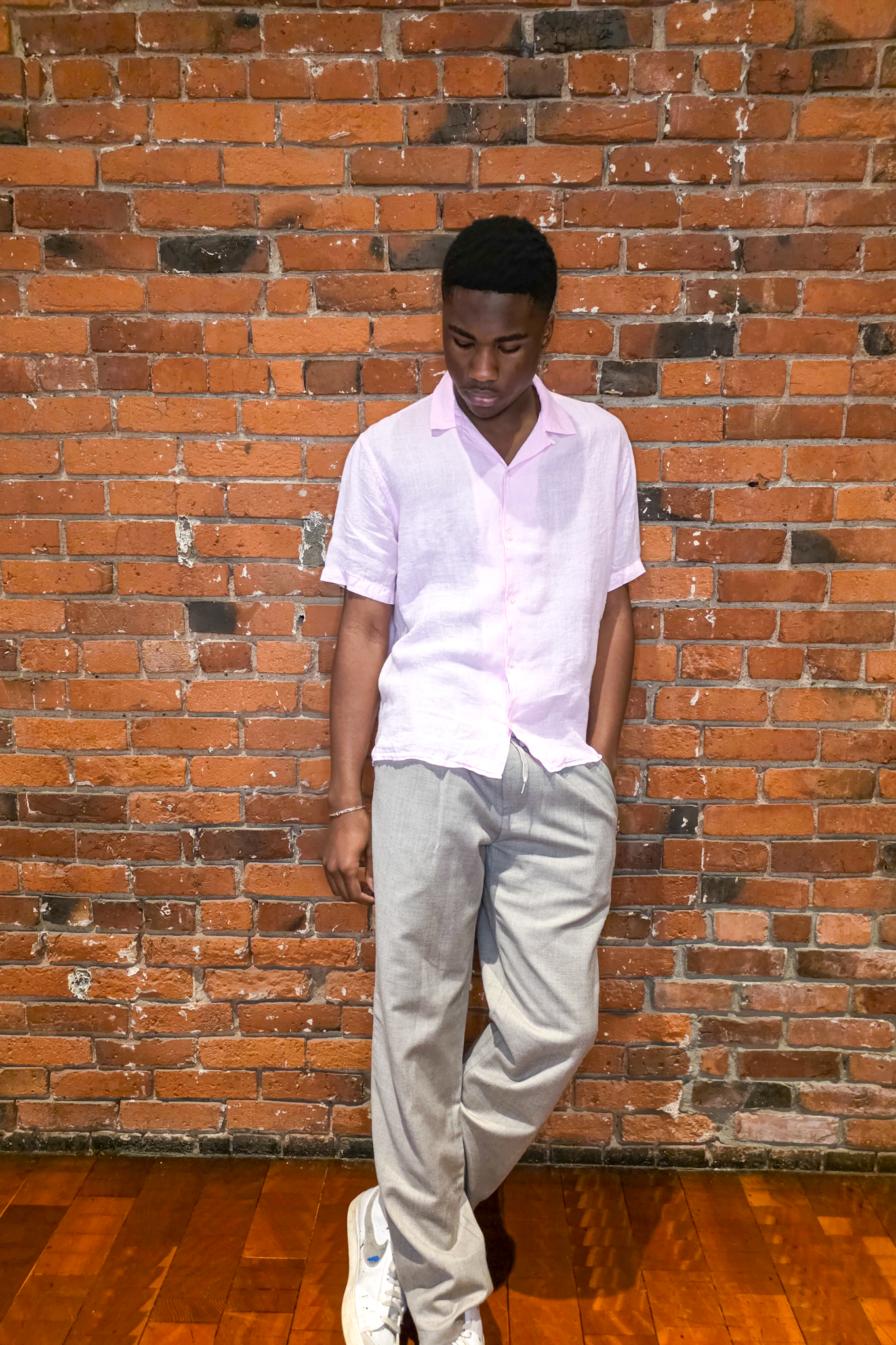 Man in a pink shirt and light gray pants standing against a brick wall.