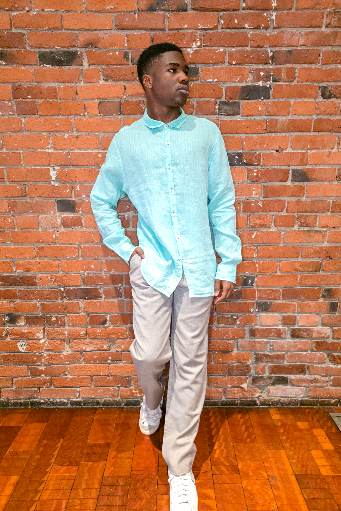 Person wearing a light blue shirt and beige pants standing against a brick wall.