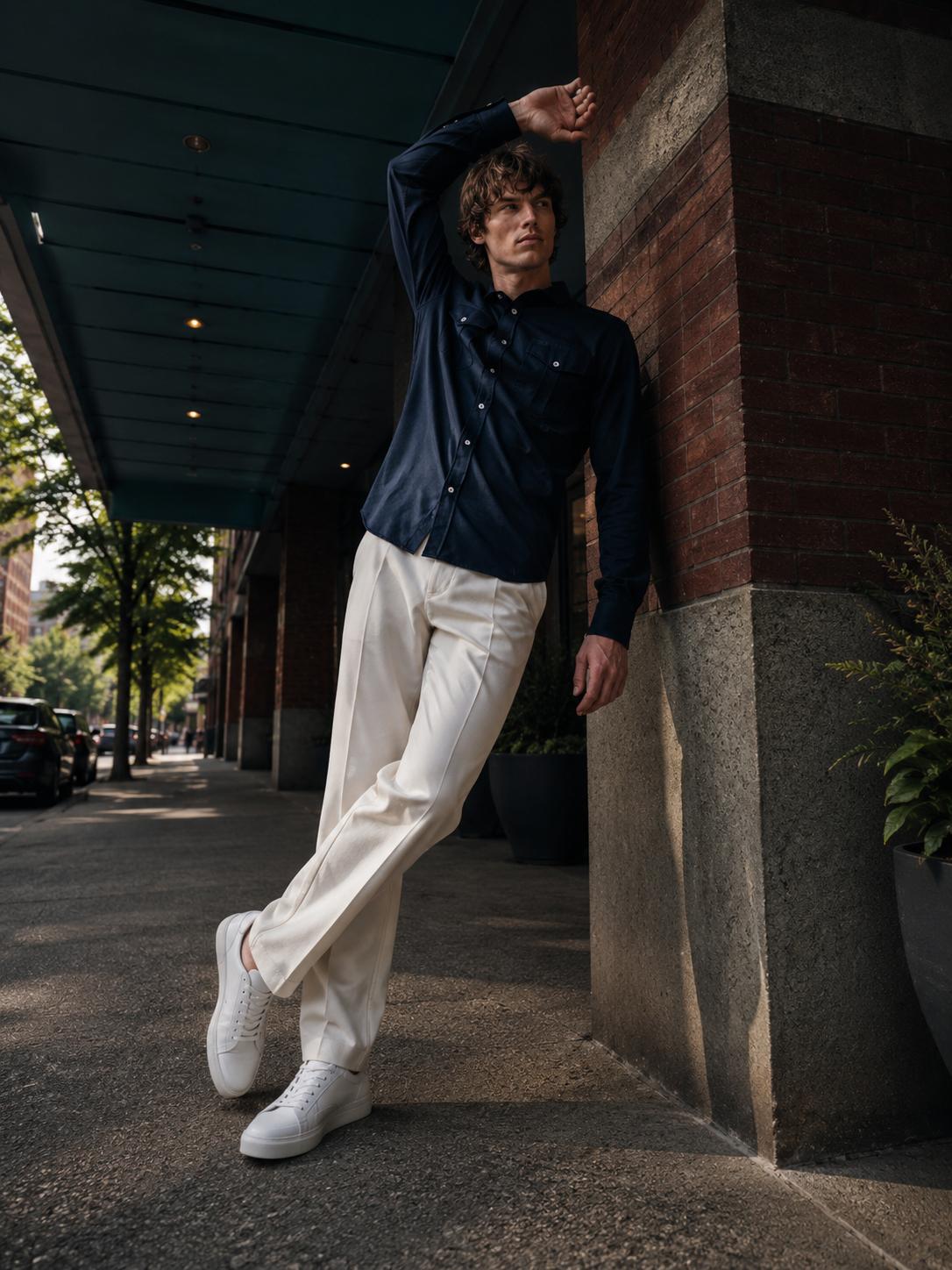 A man leans against a brick column on an outdoor urban walkway, wearing a dark navy long-sleeve button-up shirt, off-white wide-leg trousers, and white sneakers.