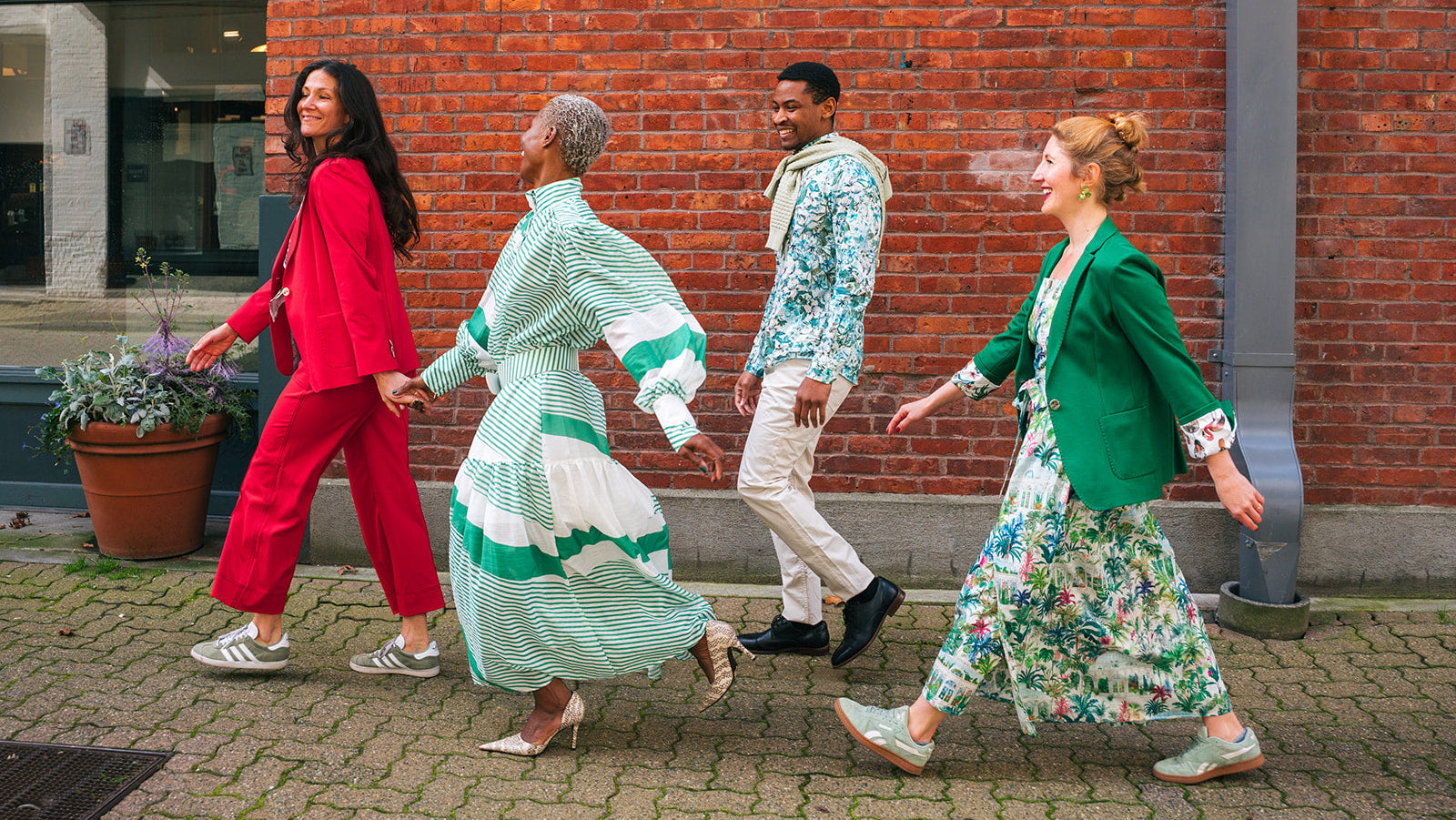 Four people walking hand in hand along a cobblestone street in front of a red brick building, each wearing a colorful, patterned outfit. The outfits feature vibrant red, green, blue, and floral prints.