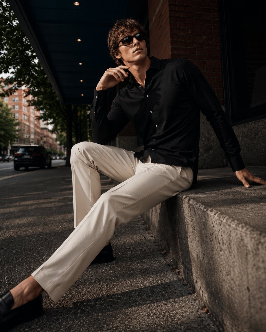 A man wearing a black long-sleeve button-up shirt and cream-colored trousers sits on concrete steps outdoors under a building overhang, wearing dark sunglasses.