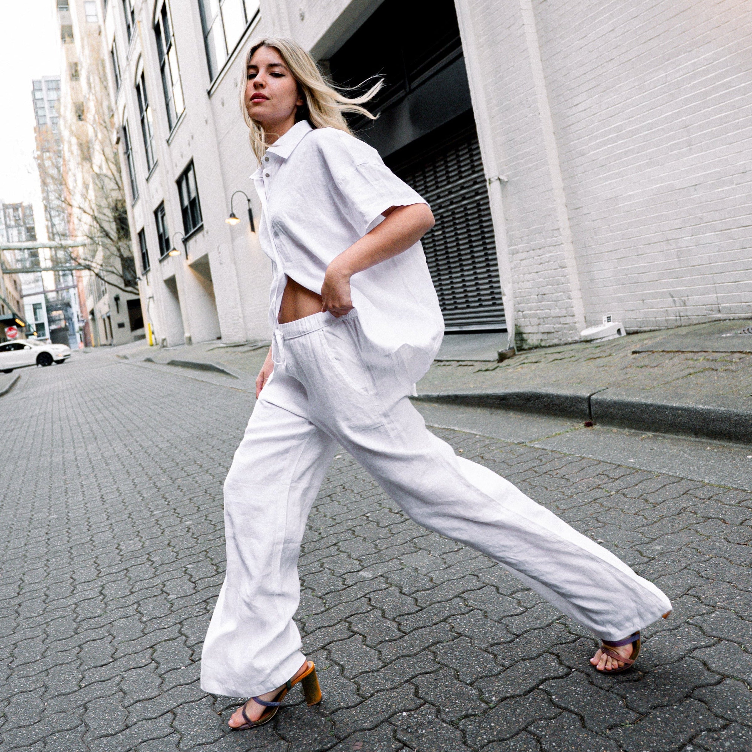 A woman in a white linen shirt and wide-leg linen pants walks across a cobblestone city street.