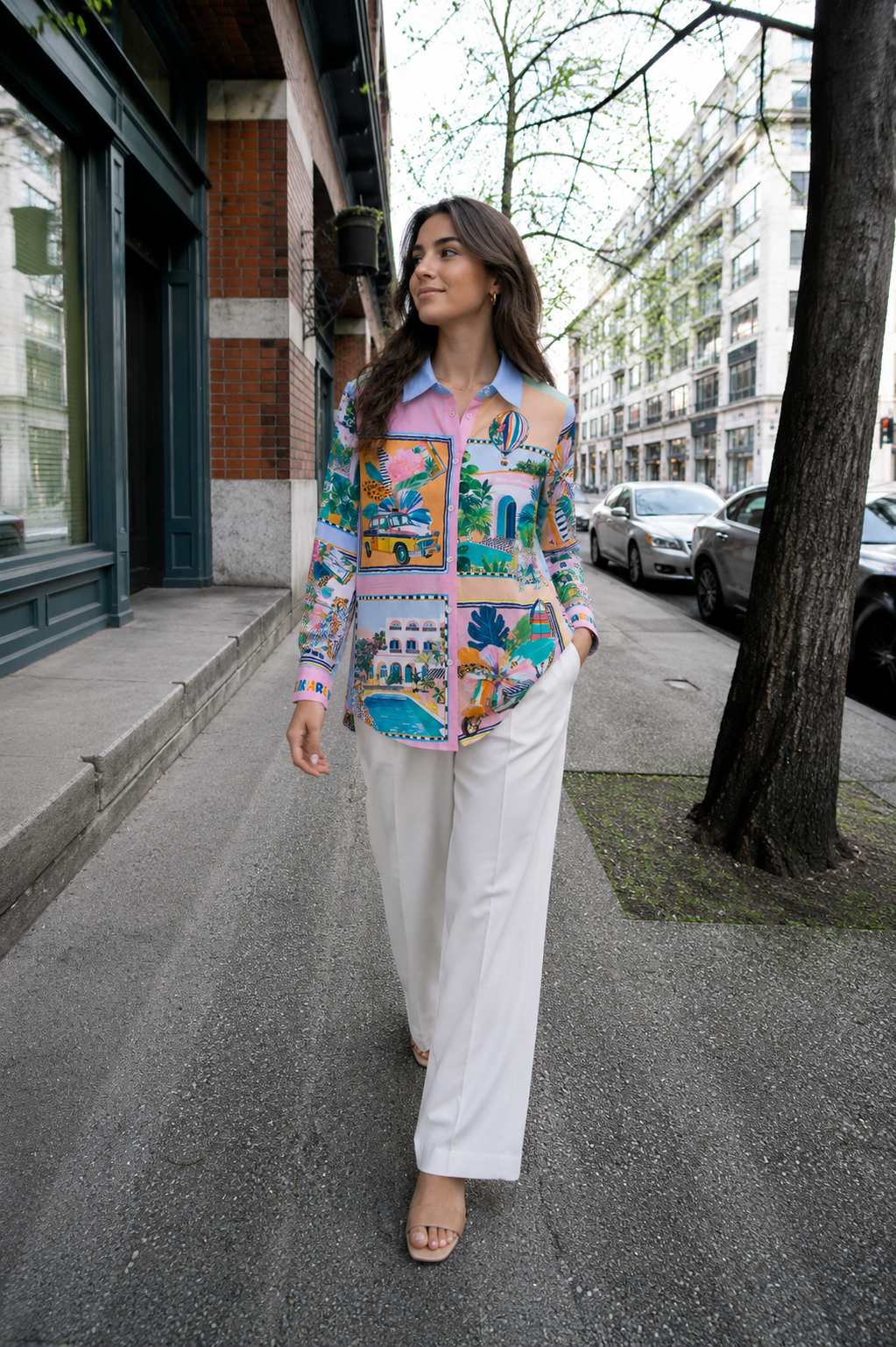 A woman walking on a city sidewalk wearing a colorful patchwork print blouse with a blue collar and white wide-leg pants.