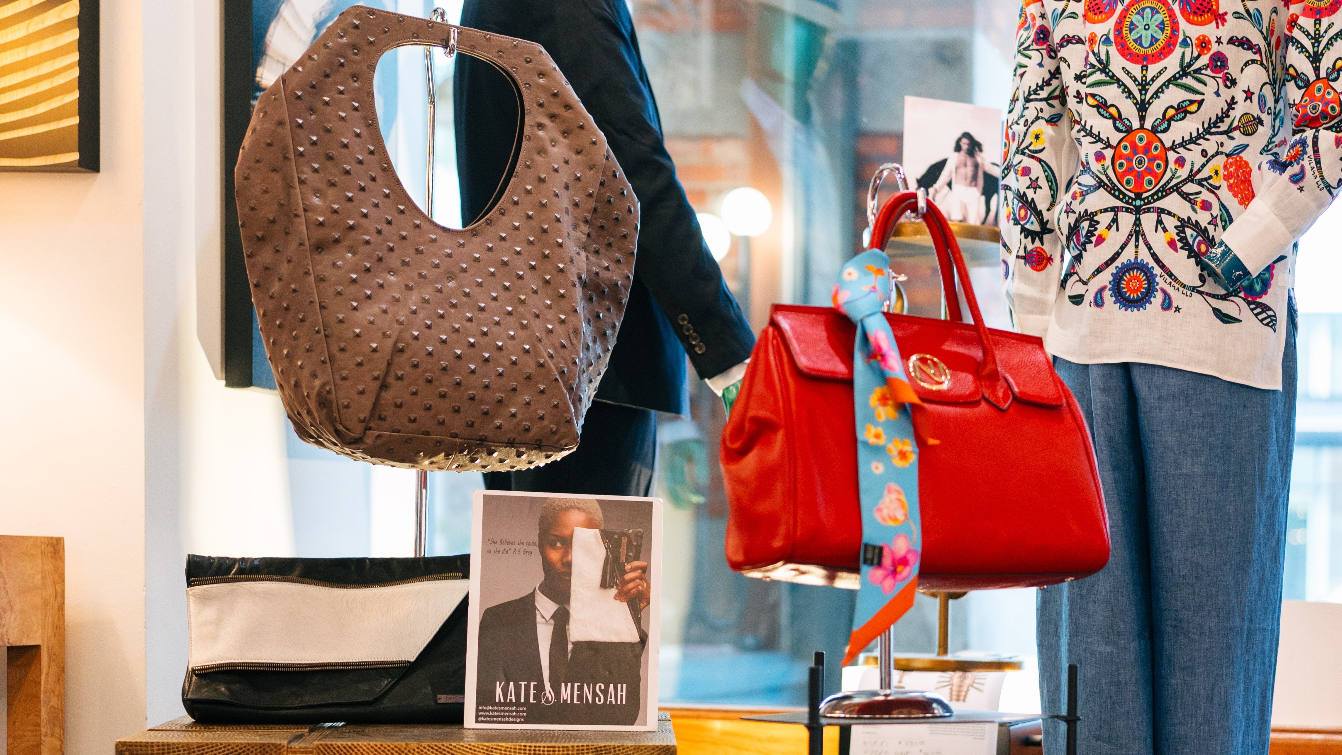 A boutique display featuring a brown woven shoulder bag hanging on the left, a bright red structured handbag with a colorful scarf strap hanging in the center, and a mannequin wearing a white embroidered sweater on the right.