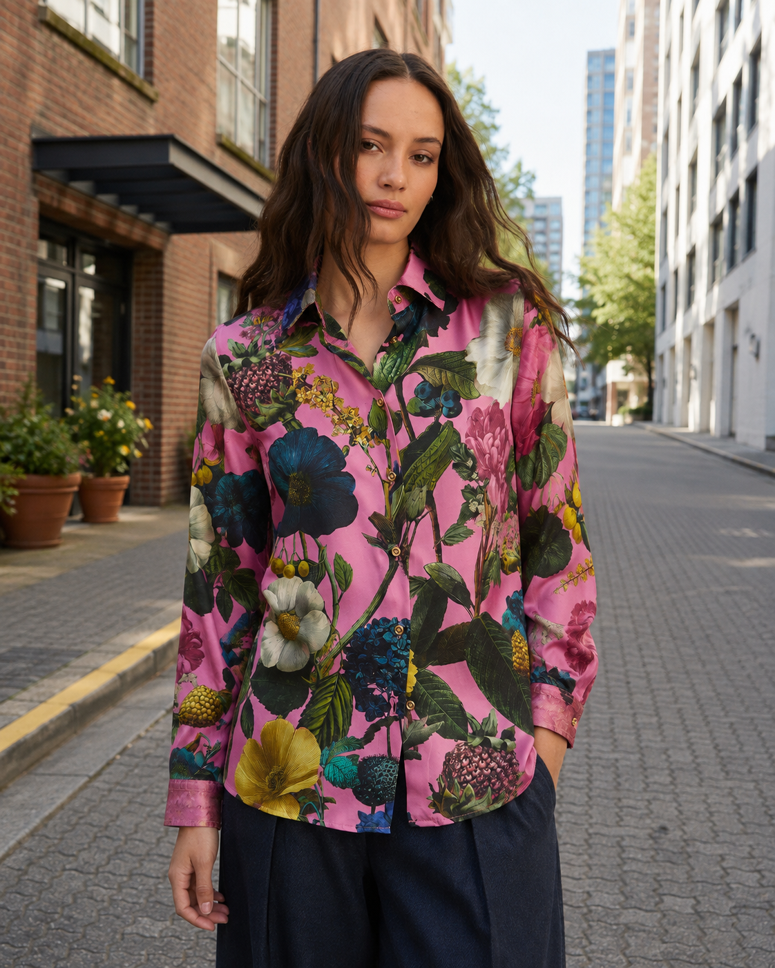 A woman stands on a city street wearing a long-sleeve button-up shirt with a vibrant pink background and a large multicolor floral print, paired with dark navy pants.